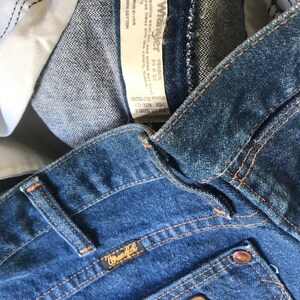 May include: A pair of blue denim jeans with a brown leather Wrangler label and a Wrangler tag sewn into the waistband. The tag includes the size 34 x 32 and the fabric content: 100% cotton.