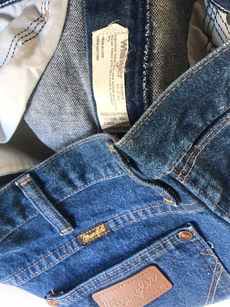 May include: A pair of blue denim jeans with a brown leather Wrangler label and a Wrangler tag sewn into the waistband. The tag includes the size 34 x 32 and the fabric content: 100% cotton.