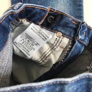 May include: Close-up of blue denim jeans with a button closure and a white tag. The tag reads "100% cotton" and includes text in English, Spanish, and French. The jeans are a slim fit.
