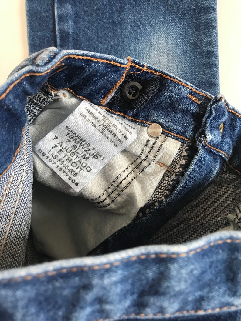 May include: Close-up of blue denim jeans with a button closure and a white tag. The tag reads "100% cotton" and includes text in English, Spanish, and French. The jeans are a slim fit.