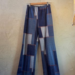 May include: Wide-leg pants with a patchwork design in shades of blue and gray, hanging on a wooden hanger. The pants feature a high waist and are made of a soft fabric. The seams are accented with a reddish-brown thread.
