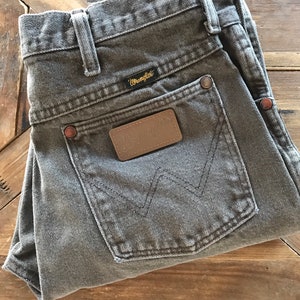 May include: A pair of gray Wrangler jeans with a brown leather patch on the back pocket. The jeans are folded in half and laid on a wooden surface.