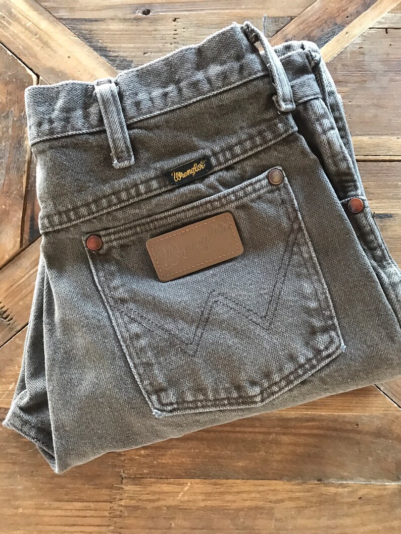 May include: A pair of gray Wrangler jeans with a brown leather patch on the back pocket. The jeans are folded in half and laid on a wooden surface.
