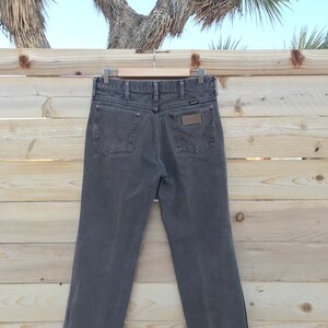 May include: A pair of dark gray denim jeans with a brown leather patch on the back pocket. The jeans are hanging on a wooden hanger against a wooden background.