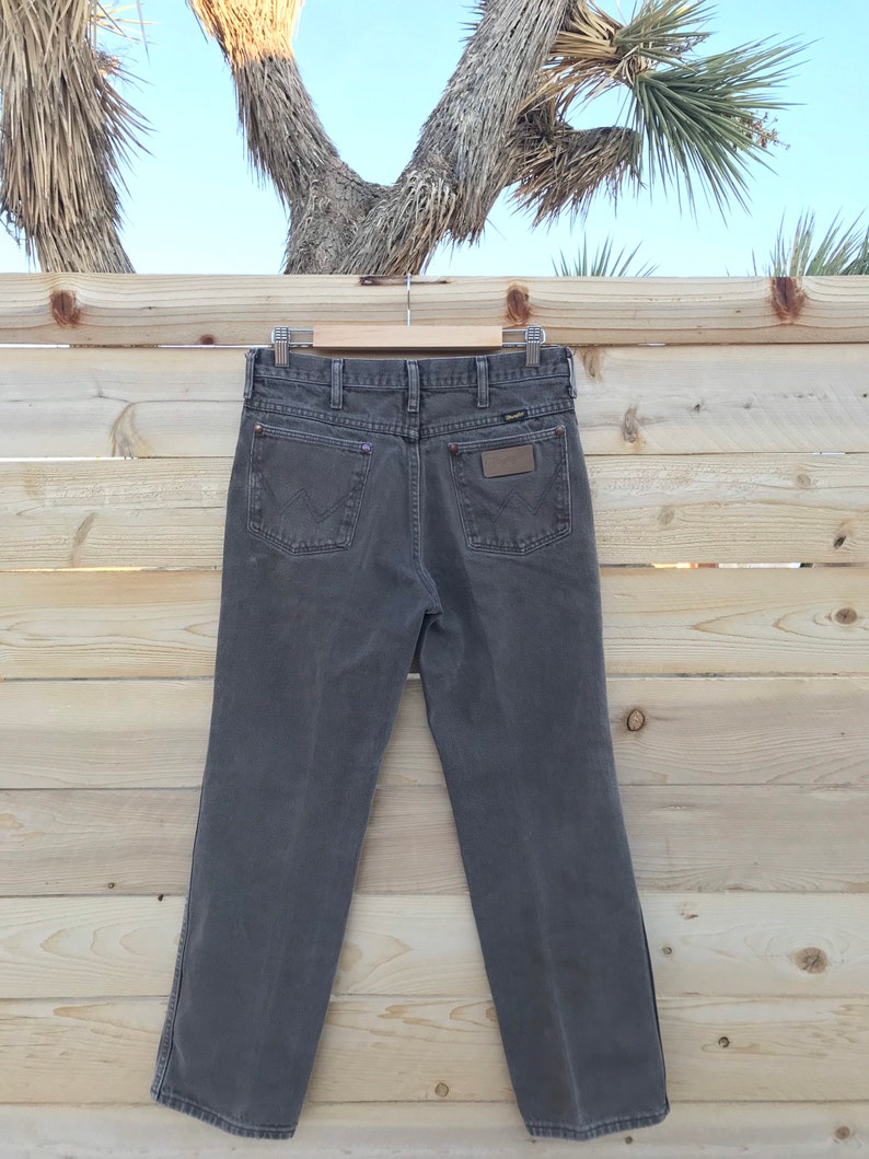 May include: A pair of dark gray denim jeans with a brown leather patch on the back pocket. The jeans are hanging on a wooden hanger against a wooden background.