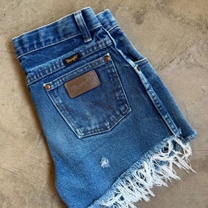 May include: Blue denim shorts featuring a high-waisted design with a Wrangler brand patch on the back pocket, copper-toned rivets, belt loops, and a frayed, raw-edge hemline. The fabric displays a medium wash with subtle distressed detailing.