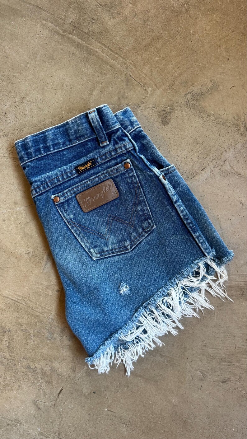 May include: Blue denim shorts featuring a high-waisted design with a Wrangler brand patch on the back pocket, copper-toned rivets, belt loops, and a frayed, raw-edge hemline. The fabric displays a medium wash with subtle distressed detailing.