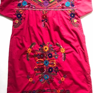 May include: A pink cotton dress with colourful floral embroidery. The dress has short sleeves and a round neckline.