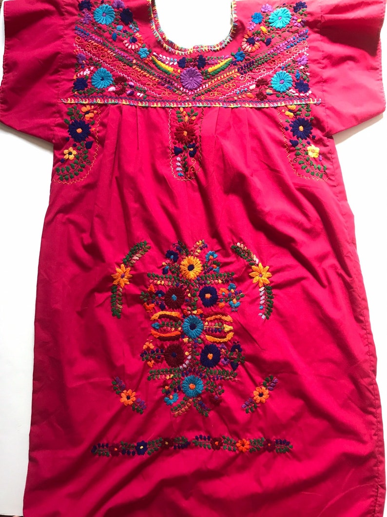 May include: A pink cotton dress with colourful floral embroidery. The dress has short sleeves and a round neckline.