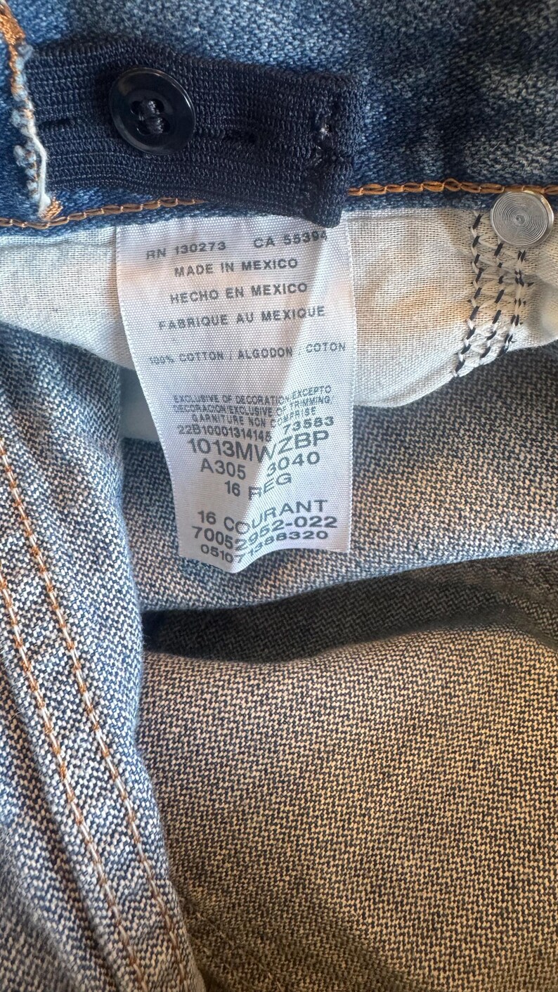 May include: Blue denim jeans featuring an internal white fabric care label with black text including RN 130273, CA 55394, Made in Mexico, 100% cotton, and size 16 REG. The jeans include a dark blue elastic waistband adjuster and a black button.