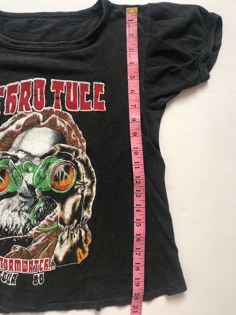 May include: Black t-shirt with a red "Jethro Tull" graphic and a cartoon illustration of a man wearing goggles. The text "Stormwatch... Tour '80" is printed below the illustration.
