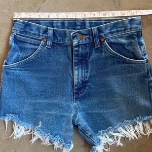 May include: Blue denim shorts with a frayed hem and a metal button closure, positioned below a white measuring tape that indicates a waist width of approximately 14 inches. The shorts feature a classic five-pocket design with copper-toned rivets.