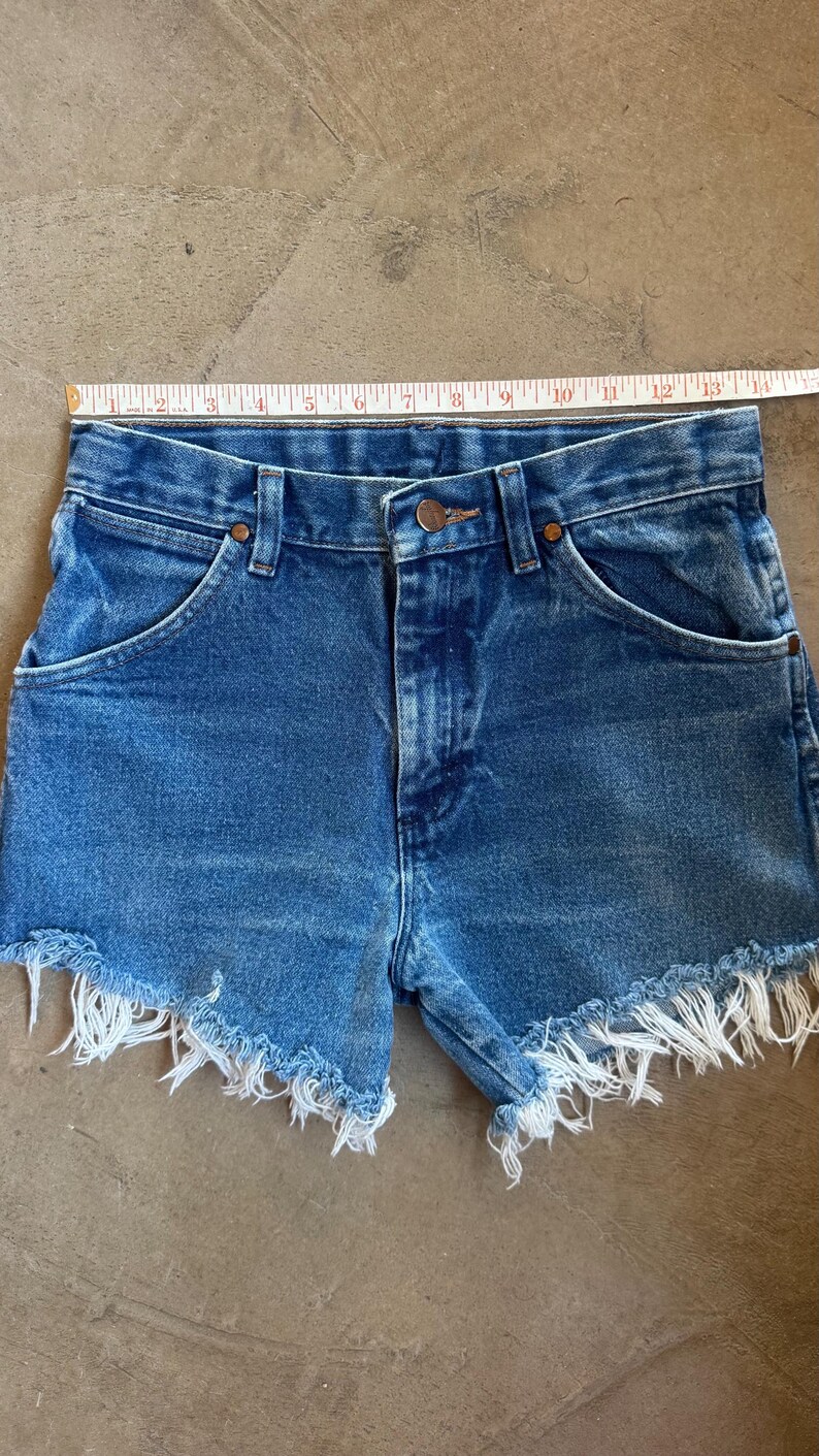 May include: Blue denim shorts with a frayed hem and a metal button closure, positioned below a white measuring tape that indicates a waist width of approximately 14 inches. The shorts feature a classic five-pocket design with copper-toned rivets.