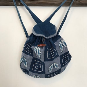Puede incluir: Una mochila de patchwork azul y gris con cierre de cord&oacute;n y un bot&oacute;n de madera. La mochila tiene un patr&oacute;n geom&eacute;trico con detalles bordados.