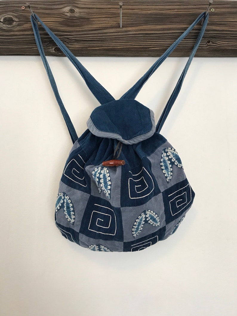 Puede incluir: Una mochila de patchwork azul y gris con cierre de cord&oacute;n y un bot&oacute;n de madera. La mochila tiene un patr&oacute;n geom&eacute;trico con detalles bordados.