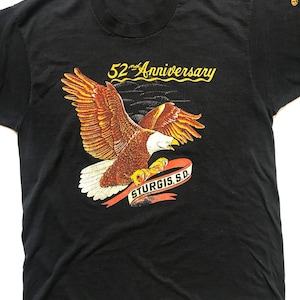 May include: Black t-shirt with a graphic of a bald eagle flying with a red ribbon that says "Sturgis, S.D." and the text "52nd Anniversary" above the eagle.