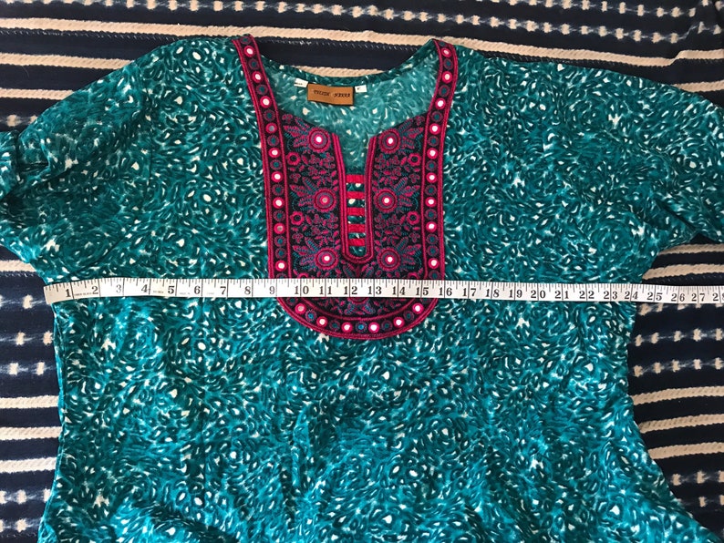 Puede incluir: Una t&uacute;nica azul turquesa con un cuello bordado en rosa y negro. El cuello tiene forma de V con una peque&ntilde;a abertura en la parte superior. La t&uacute;nica tiene un estampado de leopardo blanco y turquesa.
