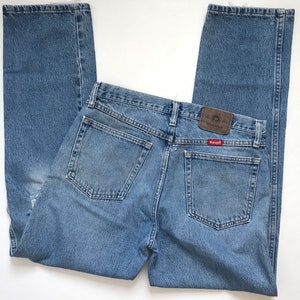 Pu&ograve; includere: Un paio di jeans in denim blu chiaro con una toppa in pelle marrone che dice "Wrangler" sulla tasca posteriore.