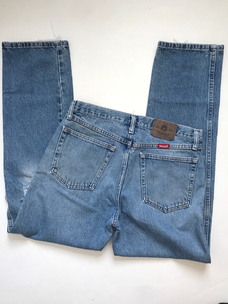Pu&ograve; includere: Un paio di jeans in denim blu chiaro con una toppa in pelle marrone che dice "Wrangler" sulla tasca posteriore.