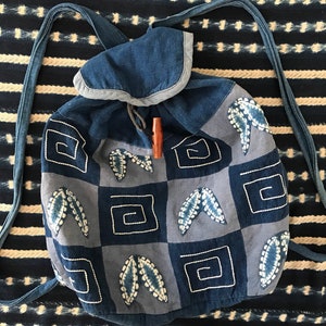 Puede incluir: Una mochila de patchwork azul y gris con cierre de cord&oacute;n y un bot&oacute;n de madera. La mochila tiene un patr&oacute;n geom&eacute;trico con hojas y cuadrados bordados.