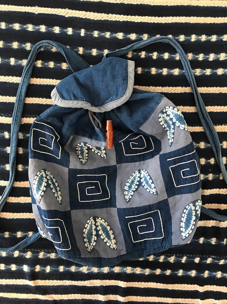 Puede incluir: Una mochila de patchwork azul y gris con cierre de cord&oacute;n y un bot&oacute;n de madera. La mochila tiene un patr&oacute;n geom&eacute;trico con hojas y cuadrados bordados.