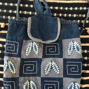Puede incluir: Una bolsa de cord&oacute;n de patchwork azul y gris con una solapa superior y correas ajustables. La bolsa est&aacute; decorada con hojas bordadas y patrones geom&eacute;tricos.