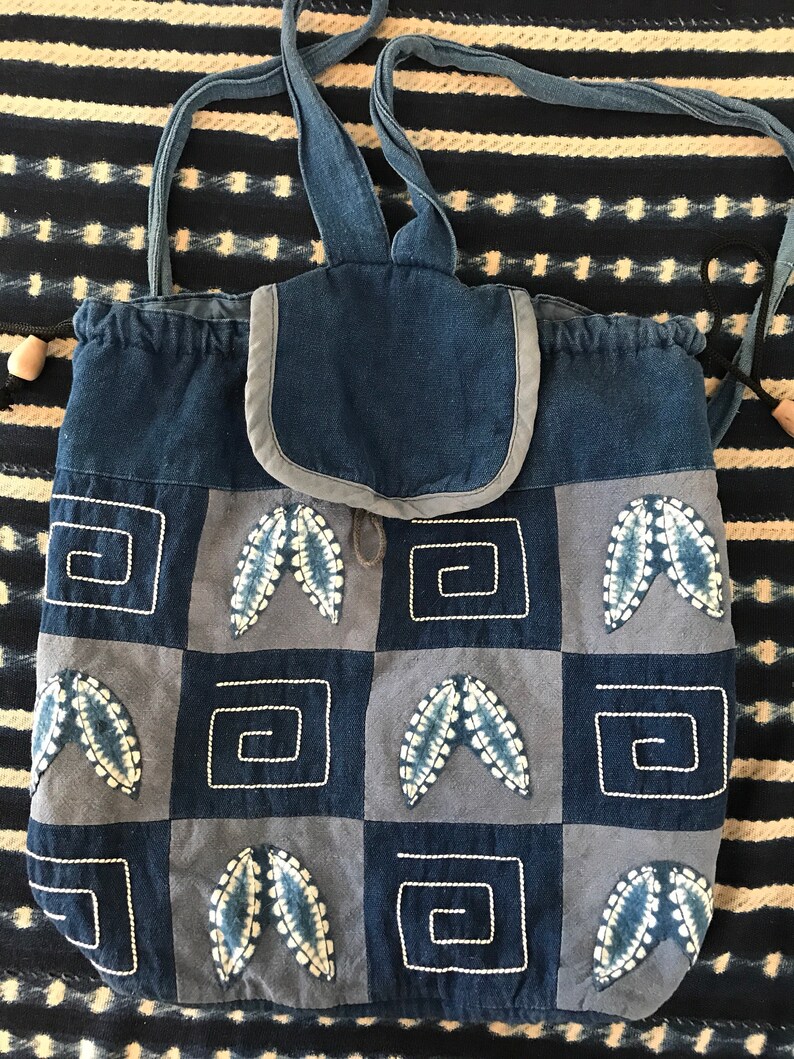 Puede incluir: Una bolsa de cord&oacute;n de patchwork azul y gris con una solapa superior y correas ajustables. La bolsa est&aacute; decorada con hojas bordadas y patrones geom&eacute;tricos.