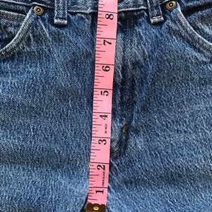 May include: A close-up of a pair of blue denim jeans with a pink measuring tape measuring the waist. The tape measures 8 inches.