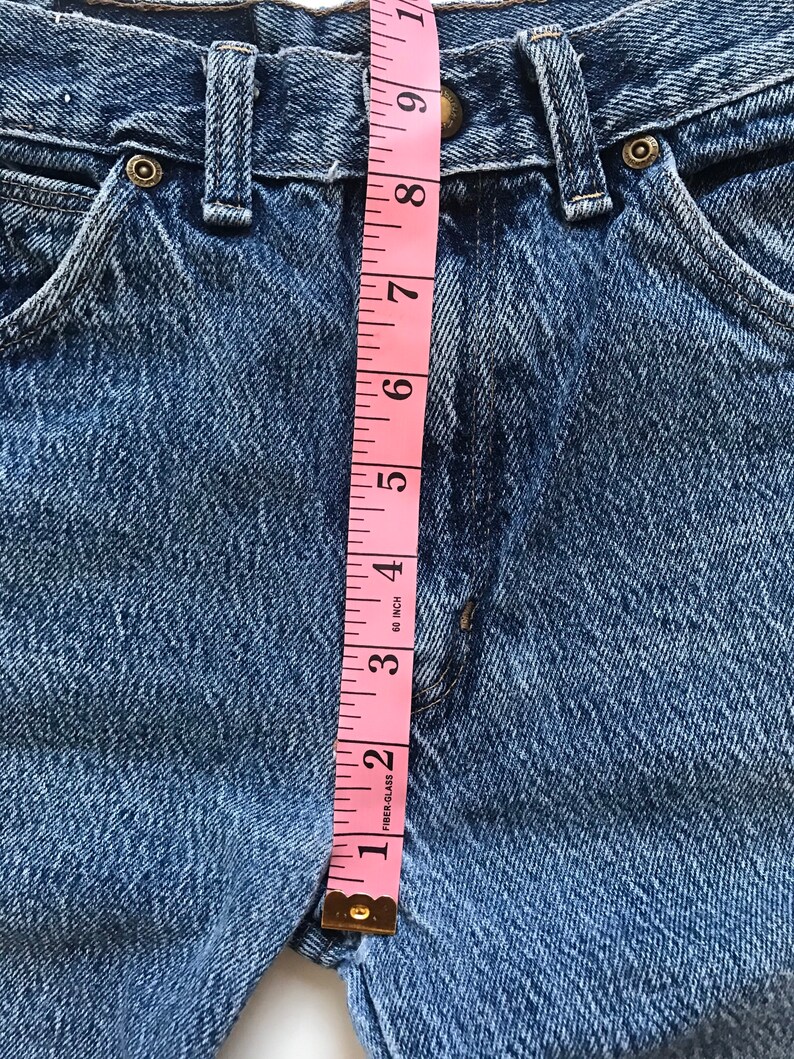 May include: A close-up of a pair of blue denim jeans with a pink measuring tape measuring the waist. The tape measures 8 inches.