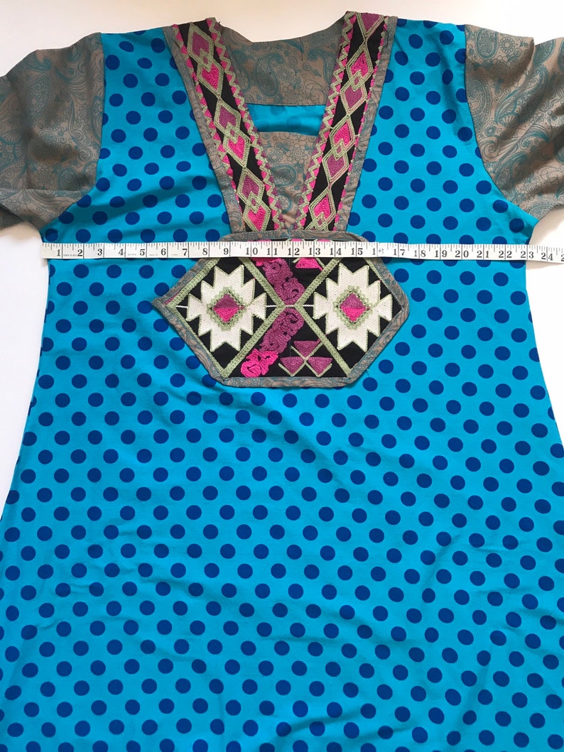 以下が含まれることがあります： 青と白のドット柄のチュニックで、カラフルな刺繍のネックラインと前面に幾何学模様が施されています。チュニックはメジャーで測定されています。