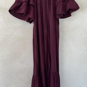 May include: A deep plum-colored midi dress featuring a V-neckline and short, layered ruffled sleeves. The garment has a loose, flowing silhouette with a gathered tier at the bottom hem, hanging from a wooden hanger against a neutral wall.