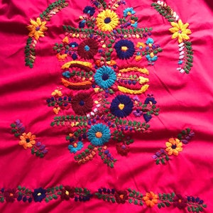 May include: A vibrant pink fabric with colourful embroidered flowers. The flowers are in shades of red, blue, yellow, green, and orange. The embroidery is detailed and intricate.