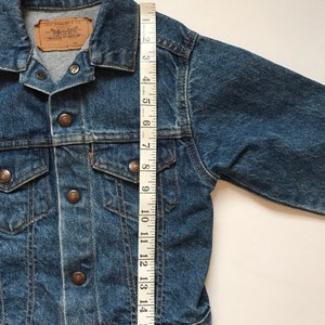K&ouml;nnte beinhalten: Eine blaue Jeansjacke mit einem Levi's-Etikett und Knopfverschl&uuml;ssen. Die Jacke wird mit einem Ma&szlig;band gemessen und zeigt eine L&auml;nge von 43 cm.