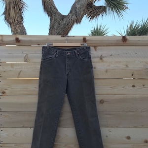 May include: A pair of dark brown denim jeans hanging on a wooden hanger against a wood panel background.