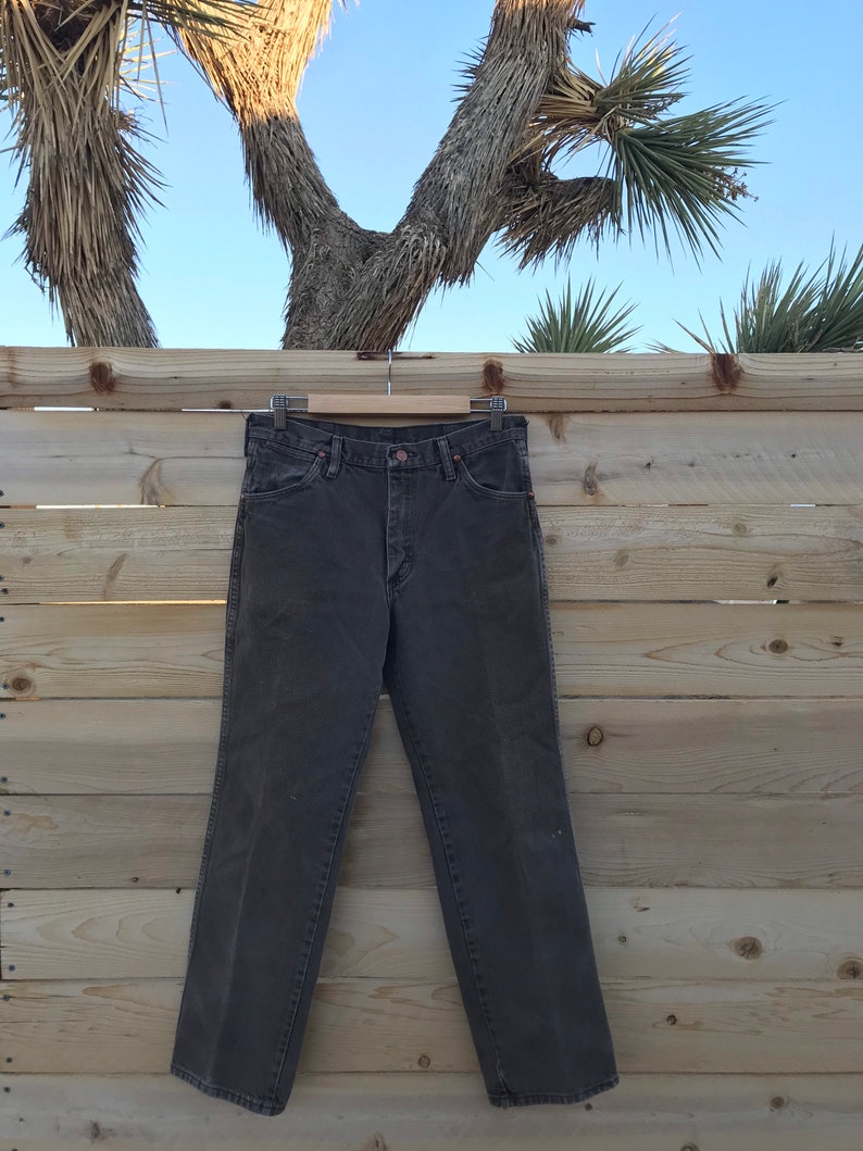 May include: A pair of dark brown denim jeans hanging on a wooden hanger against a wood panel background.