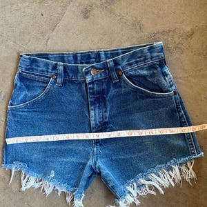 May include: Blue denim shorts with a high-waisted design, a button and zipper fly, and frayed raw-edge hems. A yellow measuring tape is placed horizontally across the front, showing a width measurement of approximately 19 inches across the waist area.