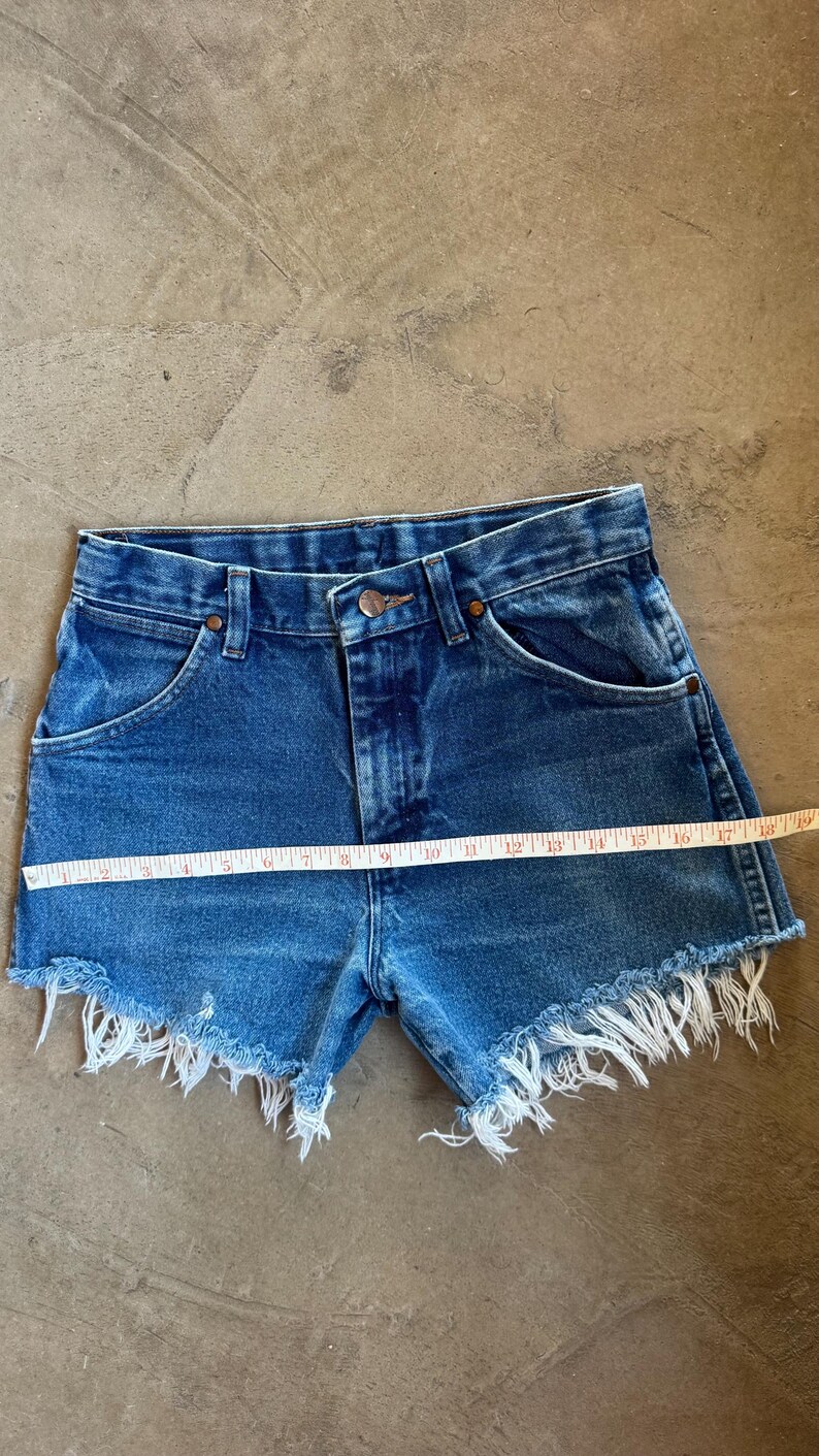 May include: Blue denim shorts with a high-waisted design, a button and zipper fly, and frayed raw-edge hems. A yellow measuring tape is placed horizontally across the front, showing a width measurement of approximately 19 inches across the waist area.