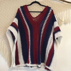 May include: A crocheted poncho with white, red, and blue stripes. The poncho has a v-neck and is made with a loose, open stitch.