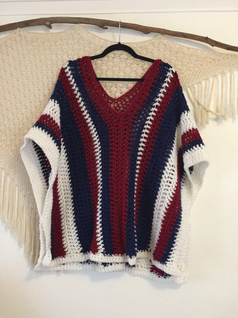 May include: A crocheted poncho with white, red, and blue stripes. The poncho has a v-neck and is made with a loose, open stitch.