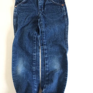 May include: Dark wash denim jeans with a classic five-pocket design. The jeans have a button and zipper closure, copper rivets, and a folded cuff at the hem. The jeans are designed for children.