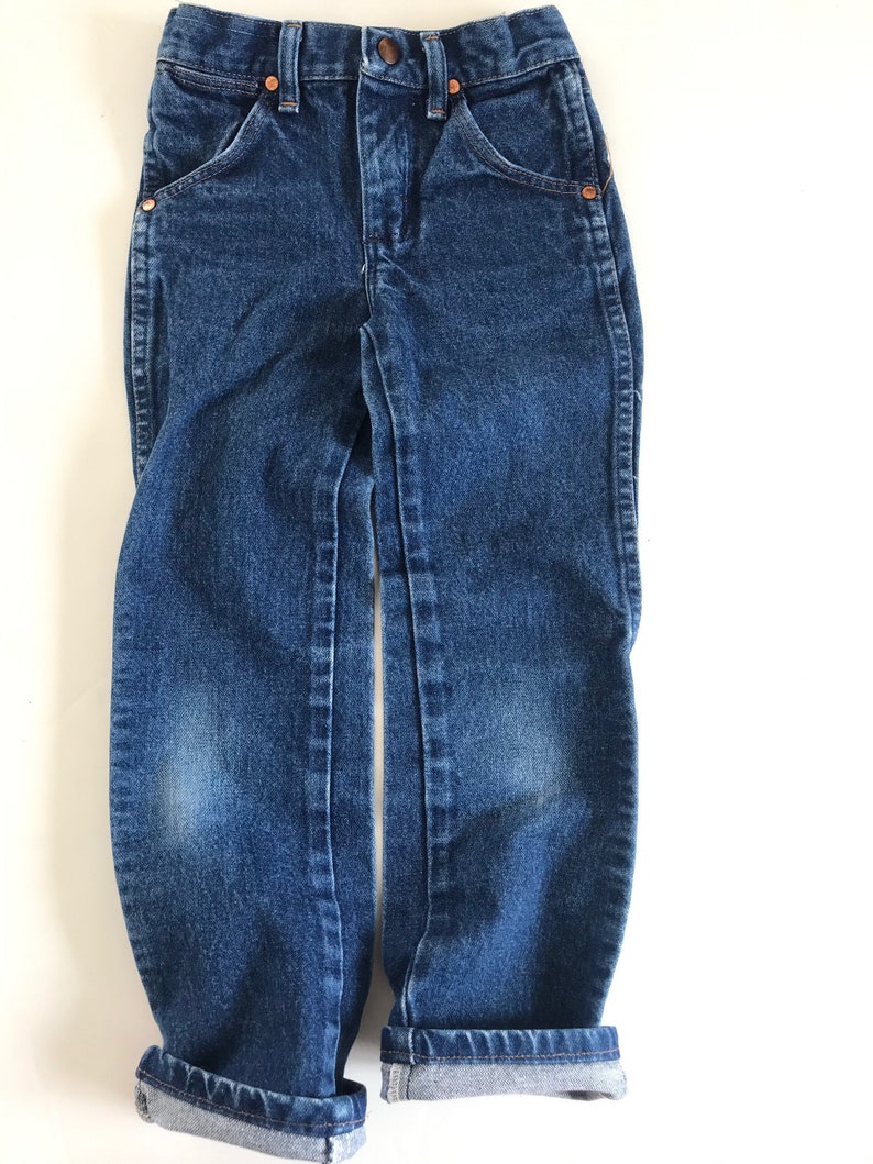 May include: Dark wash denim jeans with a classic five-pocket design. The jeans have a button and zipper closure, copper rivets, and a folded cuff at the hem. The jeans are designed for children.