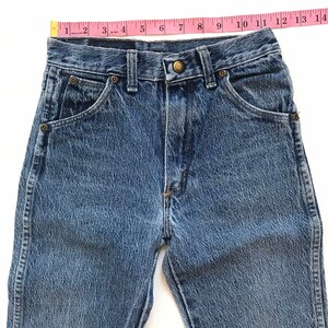 May include: A pair of light blue denim jeans with a light wash. The jeans have a classic five-pocket style and a button fly. The waist is measured with a pink tape measure.