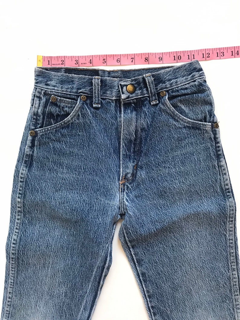May include: A pair of light blue denim jeans with a light wash. The jeans have a classic five-pocket style and a button fly. The waist is measured with a pink tape measure.