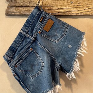 May include: Blue denim shorts featuring a frayed hem, two back pockets with a signature W-stitch pattern, copper-toned rivets, and a brown leather patch embossed with the Wrangler brand name on the back waistband area.