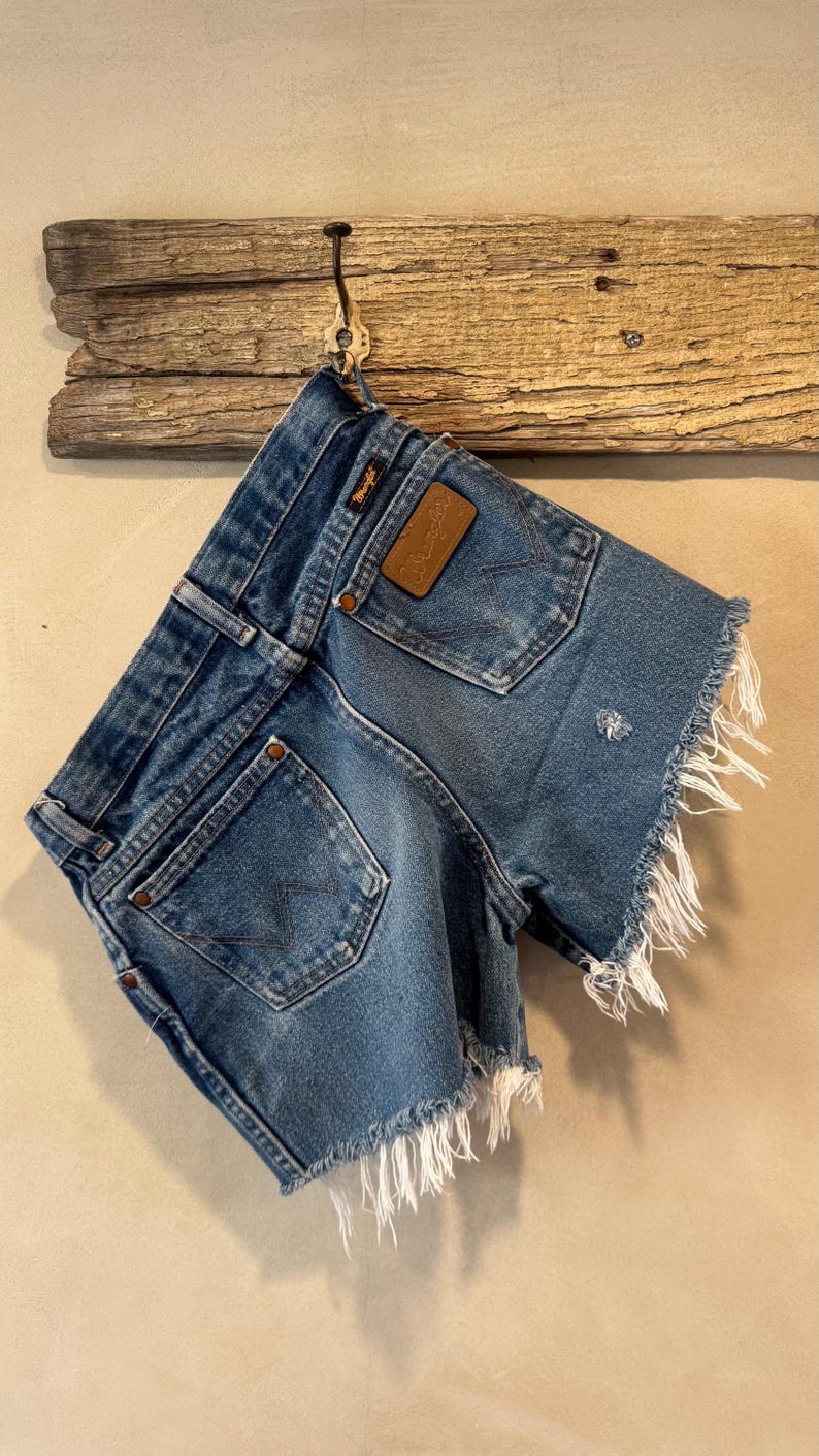 May include: Blue denim shorts featuring a frayed hem, two back pockets with a signature W-stitch pattern, copper-toned rivets, and a brown leather patch embossed with the Wrangler brand name on the back waistband area.