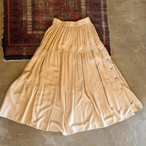 May include: A light beige, ankle-length tiered skirt featuring a gathered elastic waistband and a vertical row of small silver-toned metal buttons running down the right side, creating a subtle side slit detail in the fabric.