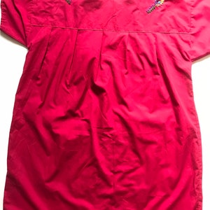 May include: A red cotton dress with embroidered floral details around the neckline. The dress has short sleeves and a loose fit.