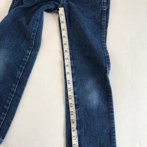 May include: Blue denim jeans with a measuring tape showing a length of approximately 20 inches. The jeans are laid flat on a white surface, showcasing the details of the fabric and stitching.