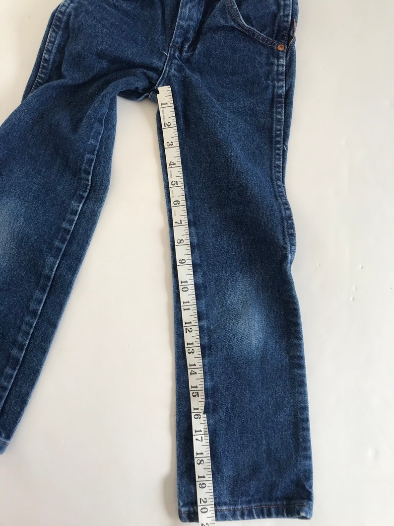 May include: Blue denim jeans with a measuring tape showing a length of approximately 20 inches. The jeans are laid flat on a white surface, showcasing the details of the fabric and stitching.