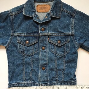 K&ouml;nnte beinhalten: Eine blaue Jeansjacke mit einem braunen Leder-Levi's-Etikett. Die Jacke hat einen Knopfverschluss und zwei Fronttaschen. Die Jacke wird mit einem Ma&szlig;band gemessen und zeigt eine L&auml;nge von 35,5 cm.